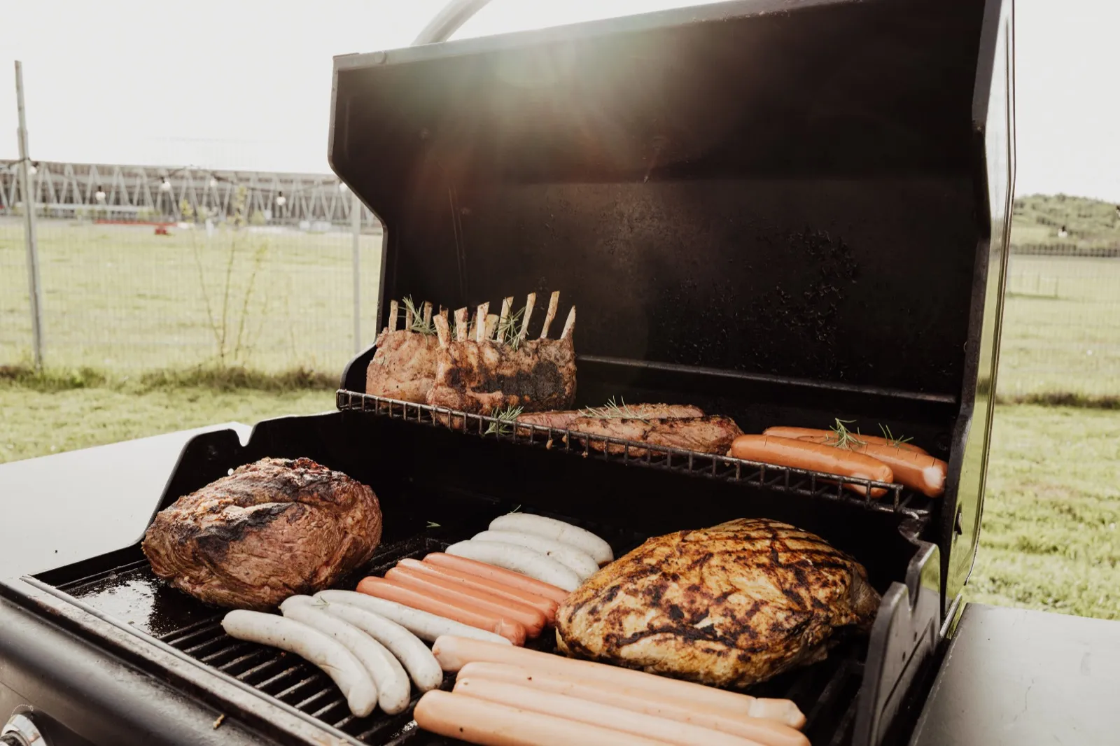 Grill mit Lammkarree, Bratwürsten und Steaks, dahinter die Wiese